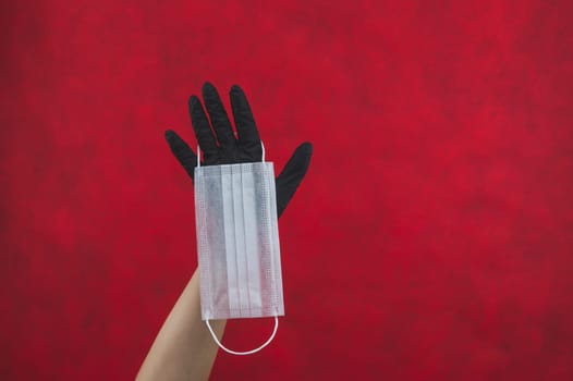 White surgical mask hangs on the hand with a black medical glove. preventive measures against respiratory illness and virus. disposable mask for protection. red background with copy space