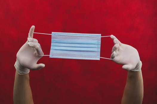 Hands in protective gloves stretch a medical mask on a red background. preventive measures against respiratory illness and virus. hands in white surgical gloves holds disposable mask for protection