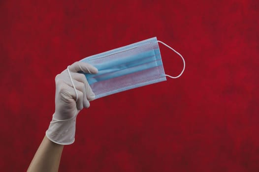 Blue medical mask in hand on a red background. preventive measures against respiratory illness and virus. hand in white surgical glove holds protective mask