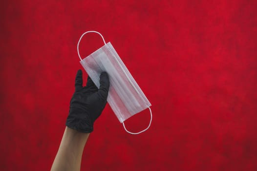 Hand in black surgical glove holds white medical mask on a red background. preventive measures against respiratory illness and virus. disposable mask for protection