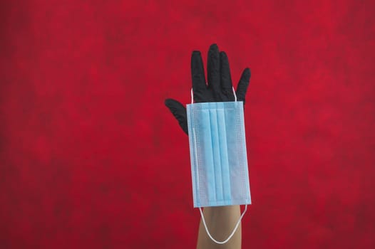 Blue medical mask hangs on the hand with a black surgical glove. preventive measures against respiratory illness and virus. red background with copy space