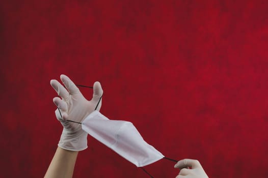 White protective mask in hands with gloves on a red background. preventive measures against respiratory illness and virus. hands in white surgical gloves holds mask for protection