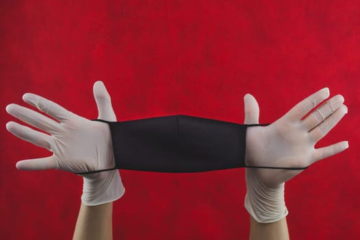 Black protective mask in hands with white surgical gloves on a red background. preventive measures against respiratory illness and virus. hands in gloves holds mask for protection