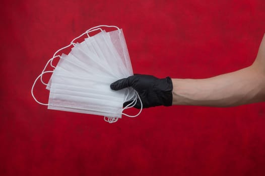 Hand in black surgical glove holds white medical masks on a red background. preventive measures against respiratory illness and virus. disposable masks for protection