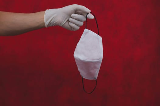White protective mask hangs on fingers. hand in white surgical glove holds medical mask on a red background. preventive measures against virus and respiratory illness. medical breathing bandage