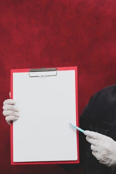 Hand in protective glove holding white blank paper sheet on red background. hand holds a pen and points out the sheet. template mock up. surface of leaflet. copy space