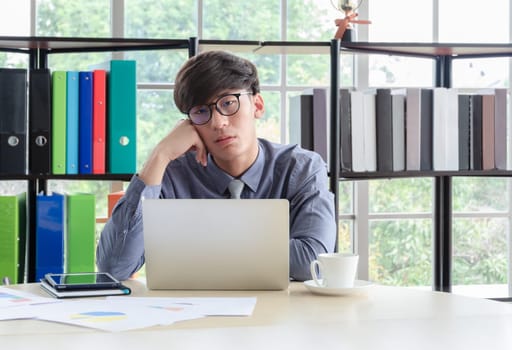 Young sleepy businessman wearing glasses sitting and holding head on hand in front of laptop computer at office desk with boried and tired face expression