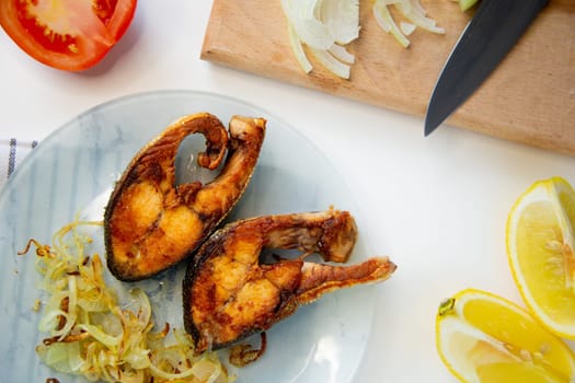 Fried fish with onions is on a plate, on a waffle towel, next to it is a cutting board with chopped onions, lemon and tomato on a white background. High quality photo