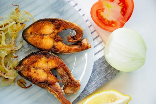 Fried fish with onions lies on a plate, on a waffle towel, next to a lemon and a tomato on a white background. High quality photo
