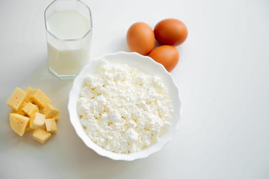 A white bowl with cottage cheese, eggs, diced cheese and a glass of milk stand on a white background. . High quality photo