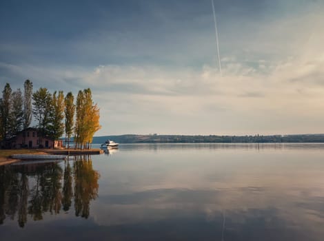 Scenic view with a house on the riverbank of Nistru river and a yacht floating. Autumn season landscape, colorful trees and beautiful sky reflected on the water surface.