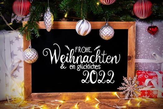 Blackboard With German Calligraphy Frohe Weihnachten Und Ein Glueckliches 2022 Means Merry Christmas And Happy 2022. Christmas Tree With Ball, Gifts And Fairy Lights