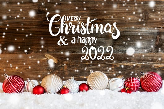 English Calligraphy Merry Christmas And A Happy 2022 On Rustic Brown Wooden Backgroud. Red And White Festive Christmas Ball Ornament On Snow And Snowflakes