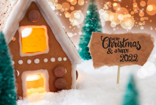 Sign With English Calligraphy Merry Christmas And A Happy 2022. Gingerbread House With Christmas Tree And Snow And Golden Sparkling Background