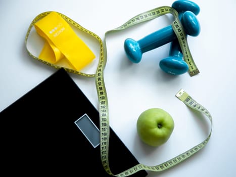 An apple, a scale, a fitness elastic band and dumbbells lie on a white background, they are entwined with a measuring tape. High quality photo