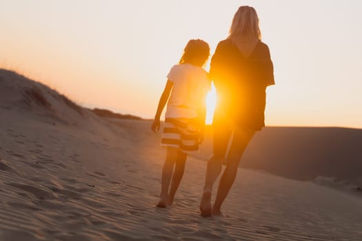 rear view of young woman and her child wearing shorts and towards the horizon on sand dunes at sunrise with sun shining between their silhouettes