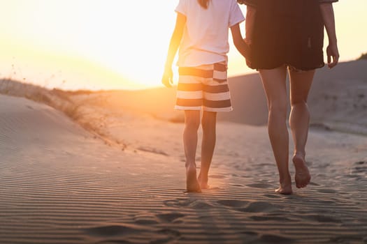 rear view of young woman and her child wearing shorts and walking holding hands towards the horizon on sand dunes at sunrise