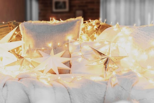 Christmas mood or New Year happy time decorations, silver paper star shape, Christmas electric light garlands on sofa with white pillows and blanket, stars decor for home interior. High quality photo