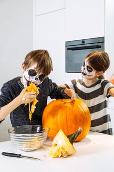 Two young children with skull face paint cutting a pumpkin in their kitchen.