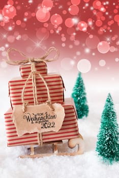 Sled With Christmas Gift And Label With German Frohe Weihnachten Und Ein Glueckliches 2022 Means Merry Christmas And A Happy 2022. Christmas Tree With Snow And Red Sparkling Background