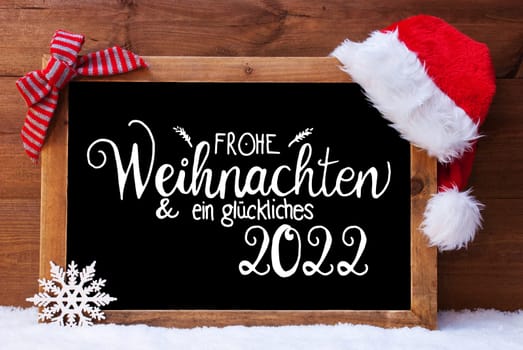 Chalkboard With German Calligraphy Frohe Weihnachten Und Ein Glueckliches 2022 Means Merry Christmas And A Happy 2022. Christmas Decoration Like Santa Hat And Bow. Wooden Background With Snow