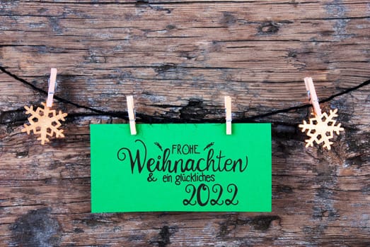 Green Label With German Calligraphy Frohe Weihnachten Und Ein Glueckliches 2022 Means Merry Christmas And Happy 2022. Rope WIth Christmas Ornamen. Rustic Wooden Background
