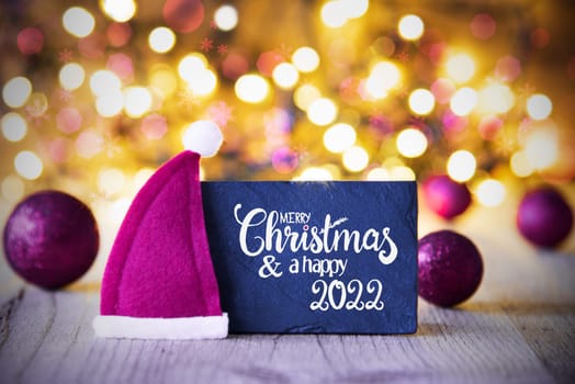 Sighn Wth English Calligraphy Merry Christmas And Happy 2022. Purple Christmas Ball Ornament With Santa Hat. Sparkling Lights Background
