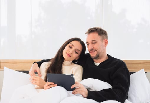 Happy young couple in sweater using a tablet PC while lying in bed at home. Lover relaxing and watching a movie together