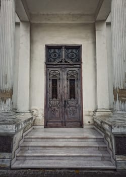 Vintage door and ornate facade details of an old historical building. Outdoors architecture elements, white columns on the doorsteps. Rustic wooden gate. The main entrance doorway