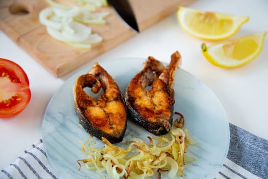Fried fish with onions is on a plate, on a waffle towel, next to it is a cutting board with chopped onions, lemon and tomato on a white background. High quality photo