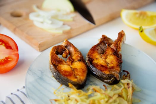 Fried fish with onions is on a plate, on a waffle towel, next to it is a cutting board with chopped onions, lemon and tomato on a white background. High quality photo