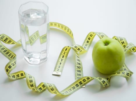 A green apple and a glass of water stand on a white background, they are entwined with a measuring tape. High quality photo