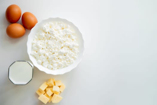 A white bowl with cottage cheese, eggs, diced cheese and a glass of milk stand on a white background. . High quality photo