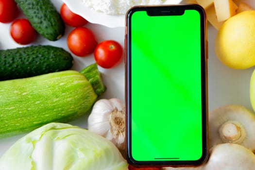 A phone with a green screen lies among fruits, vegetables and dairy products. High quality photo