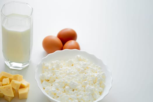 A white bowl with cottage cheese, eggs, diced cheese and a glass of milk stand on a white background. . High quality photo