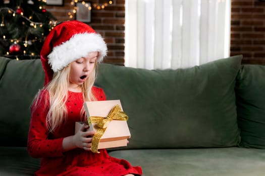 Small little shocked girl in red dress holds Christmas or New Year gift box present. Caucasian child surprised and say wow emotionally, exited because of delivered gifted surprise, expressive emotion