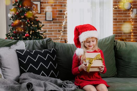 Small little smiling girl in red festive dress holding Christmas or New Year gift box present. Caucasian child dressed in Santa hat or cap sitting on green sofa in living room near decorated fir tree