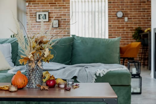 Autumn time decorated home interior. Yellow flowers bouquet in glass vase on wooden stylish table, pumpkin, red apples, leaves, candles, blanket. Green sofa, brick wall and fireplace on background