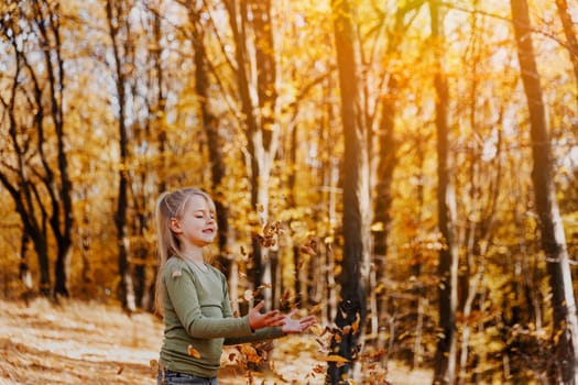 Small happy smiling little caucasian child girl playing with yellow leaves in autumn forest or garden around trees. Kid throws leaves up and they are falling down. Colorful autumn, gold colors around