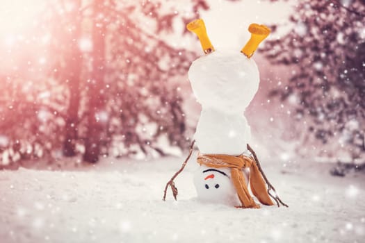 Cheerful snowman with orange scarf and in yellow boots stands upside down in winter forest. Panoramic view of the beautiful background in snowy natural park in snowfall.