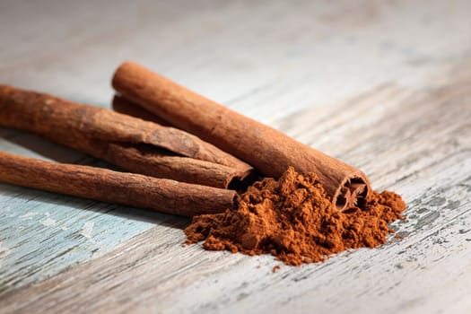 Close-up of several rolled cinnamon sticks with cinnamon powder lying in front of them on a wooden surface with paint residues.