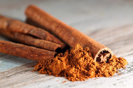 Close-up of several rolled cinnamon sticks with cinnamon powder lying in front of them on a wooden surface with paint residues.