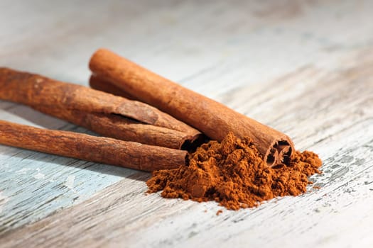 Close-up of several rolled cinnamon pieces with cinnamon powder lying in front of them on a wooden surface with paint residues.