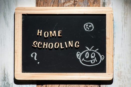 Frontal close-up of a small black chalk board made of wooden letters with the words Home Schooling and a stick figure drawing made of chalk on a wooden surface in the harsh sidelight.