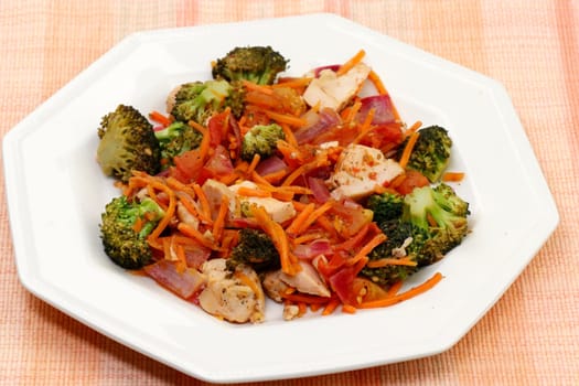 Prepared meal of chicken meat with broccoli, carrots, purple onion and oregano closeup. Delicious lunch of chicken bites with vegetables and seasonings on a white octagon plate on a place mat.