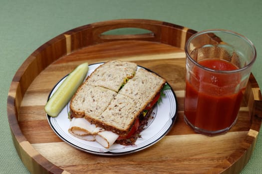 On a round wood tray on a plate is a turkey cold cut sandwich with tomato, lettuce and mustard near a vegetable beverage. Turkey and vegetable sandwich with a dill spear pickle and tomato drink meal.