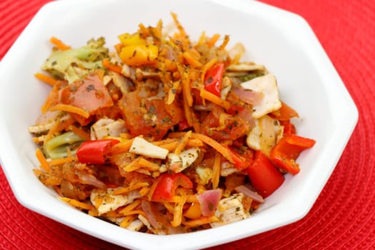 In an octagon bowl is a prepared lunch of organic turkey meat and a variety of vegetables close-up on a red place mat. Cooked meal of turkey cold cuts sweet bell pepper and other vegetables and herbs