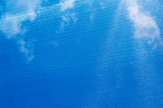 Blue sunshade that you can see sunlight and white clouds above viewer that filters bright sunshine. Large flat blue sun shade textile structure that you can see through that reduces the sunshine below.