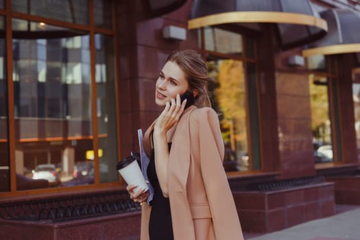 A busy woman with a newspaper, laptop and takeaway coffee in one hand is walking down the street and talking on the phone. Big city rhythm, rush