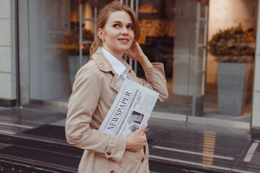 Beautiful girl in coat with newspaper and smiling while walking outdoors